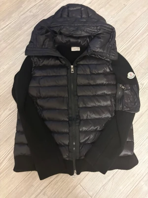 Svart Moncler pufferjacka med stickade ärmar - Svart pufferjacka från Moncler med stickade ärmar och huva. Jackan har quiltad front i glansig polyester, dragkedja med Moncler-logga och patch på ärmen. Snygg kombination av puffer och stickat, perfekt för kalla dagar.