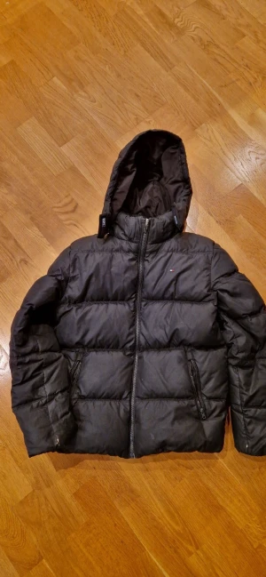 Tommy Hilfiger svart dunjacka stl 152 - Svart dunjacka med dragkedja och huva. Jackan har flera fickor med dragkedja och en liten Tommy Hilfiger-logga. Finns även Tommy Hilfiger varumärket uppe vid huvan. Små hål vid ärmslutens undersida samt litet hål på framsidans nederkant (samt någon fläck som gör tyget aningens glansigare). Se bilder. Från rök- och djurfritt hem.