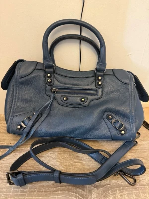 Blå Citybag likande väska/handväska - Snygg blå  Citybag likande väska/handväska i mjukt skinn med klassiska silverdetaljer och dragkedjor. Väskan har både handtag och avtagbar axelrem, samt coola dekorativa spännen och långa läderband. Perfekt storlek för dina essentials och har flera fack med dragkedja.