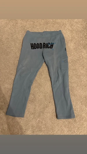 Blåa Hoodrich mjukisbyxor med ficka - Snygga blå mjukisbyxor från Hoodrich med svart logga bak och en praktisk ficka på sidan av benet. Byxorna har en slim passform och är perfekta för en chill och avslappnad stil. Tillverkade i mjukt material för maximal komfort.