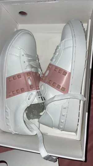 Valentino Rockstud sneakers vit/rosa - Säljer ett par Valentino Rockstud sneakers i vitt skinn med breda ljusrosa band och ikoniska nitar över ovandelen. Skorna har vita snören,och säljer dessa för endast 750