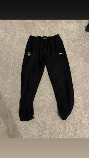 Svarta Jordan PSG mjukisbyxor - Svarta mjukisbyxor från Jordan och PSG med gula loggor på framsidan. Byxorna har resår i midjan och vid bensluten för en skön passform. Perfekta för chill eller sport. Materialet är mjukt och bekvämt.