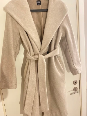 Beige kappa med huva från Zara - Snygg beige kappa från Zara med stor huva och bälte i midjan. Kappan har lång ärm, bred krage och är tillverkad i ett mjukt ullblandat material. Perfekt för dig som vill ha en stilren och mysig look under höst och vinter.