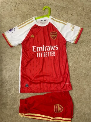 Arsenal matchställ G.Jesus nr 9 - Säljer ett Arsenal matchställ med tröja och shorts, officiell Adidas med G.Jesus och nummer 9 på ryggen. Röd och vit med guldiga detaljer och klubbmärke på både tröja och shorts. Perfekt för unga fotbollsfans som vill köra stilen på planen.