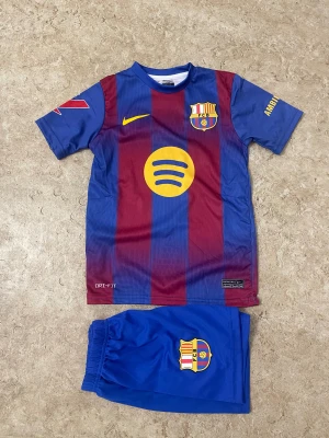 FC Barcelona Raphinha matchställ Nike - Säljer ett FC Barcelona matchställ med Raphinha 11-tryck. Tröjan har klassiska blå och vinröda ränder, gula detaljer och klubbmärke på bröstet. Korta ärmar, DRI-FIT-material och tillhörande blå shorts med klubbmärke. Perfekt för fotbollsälskare!