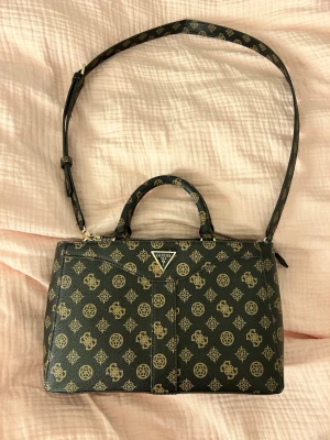Mörkbrun handväska från Guess - Snygg mörkbrun handväska från Guess med guldiga logomönster över hela väskan. Väskan har både handtag och justerbar axelrem samt flera fack med dragkedja. Tillverkad i syntetmaterial med klassisk Guess-triangel framtill.
