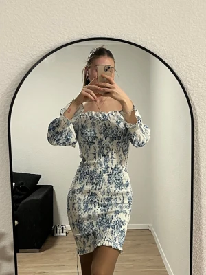 Blommig kortklänning med puffärm - Säljer en superfin vit och blå kortklänning med blommigt mönster och puffiga långärmar. Klänningen har smockad midja och axelparti, samt en söt volangkant längst ner. Perfekt för dig som gillar romantisk och trendig stil.