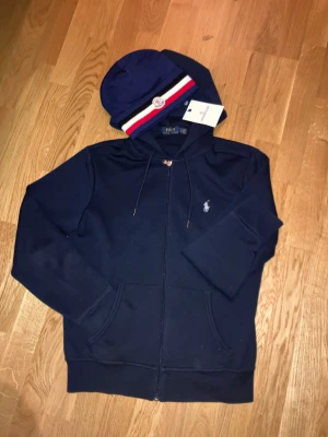 Zip up och mössa moncler-Ralph lauren - Snygg mörkblå hoodie från Polo Ralph Lauren med dragkedja, huva med dragsko och klassisk broderad logga på bröstet-Snygg moncler mössa skönt material och perfekt till dailywear. Perfekt för en clean och chill outfit. Både (mössan och zippern) sista bilden är för inspiration, pris går att diskutera vid snabb affär.