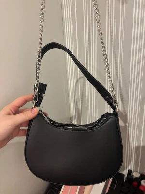 Svart axelväska med kedja - Snygg svart axelväska i skinnimitation med silverfärgad kedja som axelrem och dragkedja upptill. Väskan har en rundad form och minimalistisk design, perfekt för att lyfta din outfit. Enkel att matcha och rymmer det viktigaste du behöver ha med dig.