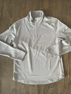 Grå golftröja från Next Tee - Snygg grå långärmad tröja från Next Tee Golf Clothes med half zip och hög krage. Materialet är lätt och känns sportigt, perfekt för golf eller chill. Diskret logga på bröstet och stilren design som funkar till många outfits.