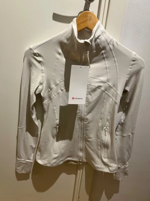 Lululemon Jacket  - Snygg vit träningsjacka från Lululemon med figurnära passform, hög krage och dragkedja framtill. Jackan har praktiska fickor med dragkedja och coola sömdetaljer. Materialet är mjukt och stretchigt polyester, perfekt för aktiva dagar.