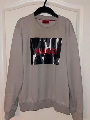 Beige sweatshirt från Hugo med tryck - Snygg beige sweatshirt från Hugo med ett coolt svartvitt mönster och röd logga på bröstet. Tröjan har rund hals, ribbade muddar och lång ärm. Perfekt för dig som gillar streetstyle och vill sticka ut med ett grafiskt tryck.