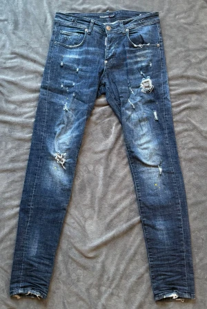 Mörkblåa Dsquared2 ICON jeans med slitningar  - Säljer ett par blå Dsquared2 ICON. Jeansen har slitna detaljer, hål och färgstänk för en edgy look. 