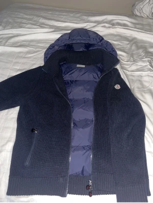 Mörkblå dunjacka från Moncler - Snygg mörkblå dunjacka från Moncler med stickade ärmar och quiltad kropp. Jackan har huva, dragkedja framtill och två fickor med dragkedja. Perfekt för kyliga dagar och har en clean, sportig look.