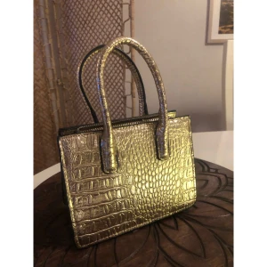 Guldig handväska med krokodilmönster - Snygg liten handväska i guld med krokodilmönstrad yta och två stadiga handtag. Väskan har en rektangulär form och stängs med dragkedja upptill. Perfekt för att lyfta din outfit med lite extra bling och cool struktur. Använd 1 gång. Väskan är från H&M.