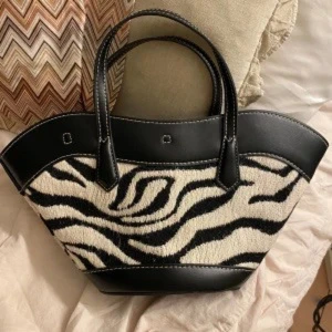 Zebramönstrad handväska - Snygg handväska med zebramönster i svart och vitt, kombinerat med svarta skinnpartier och kontrastsömmar. Väskan har två handtag och en unik, bred form som sticker ut. Perfekt för dig som vill ha en statement-accessoar med cool vibe.