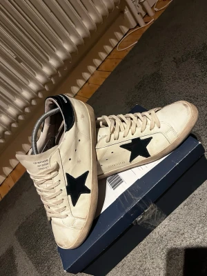 Golden Goose vita sneakers med svart stjärna - Golden Goose sneakers i vitt skinn med svart stjärna på sidan och svart häl. Klassisk rund tå, platt sula och snörning framtill. Snygg kontrast mellan den ljusa ovandelen och den svarta detaljen. Perfekt för dig som gillar streetstyle och unika detaljer.