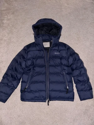 Mörkblå jacka från GANT med huva - Helt ny och oanvänd! Riktigt snygg mörkblå jacka från GANT med huva och dragkedja. Jackan har två snedställda fickor med dragkedja, broderad GANT-logga på bröstet och rund patch på ärmen. 