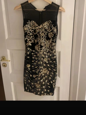 Svart och guld glittrig festklänning - Supersnygg svart kort klänning med guldiga paljetter och broderier i blommigt mönster. Klänningen är ärmlös med mesh upptill och har dragkedja i ryggen. Perfekt för dig som vill sticka ut med en glamorös look.