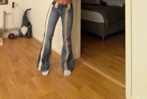 Vintege blå,  beige bootcut jeans med ränder. - Unika blå jeans med breda beige ränder längs benen och bootcut passform. Byxorna har låg midja, bälteshällor och dekorativa knappar framtill. Materialet är klassisk jeans och designen är riktigt retro med coola kontraster. Dessa jeans är verkligen super coola, jag säljer dem då dem inte används så mycket, jag hoppas dem kommer till någon som kan uppskatta dem mer! Dem är köpta i London för ca 1år sedan. Jag är 159cm lång för referens, jag skulle ändå vilja påstå att dem är små i storleken!🥰