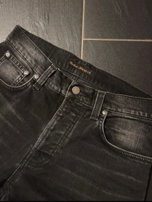 Svarta slim fit jeans från Nudie Jeans - Snygga svarta jeans från Nudie Jeans med slim passform och klassisk femficksdesign. Jeansen har slitna detaljer och subtila tvättade partier som ger en cool look. Materialet är jeans och bakfickorna har Nudie Jeans signaturbroderi.