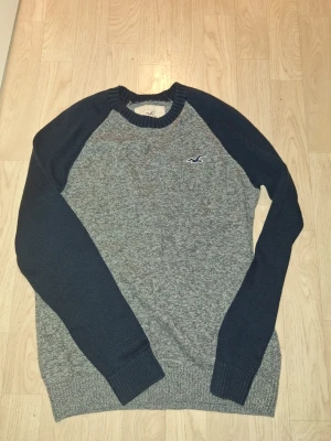 Grå och marinblå stickad tröja Hollister - Snygg stickad tröja från Hollister i grått med marinblå ärmar och ribbade muddar. Klassisk crewneck med Hollister-logga broderad på bröstet. Perfekt för dig som gillar en enkel men stilren look.