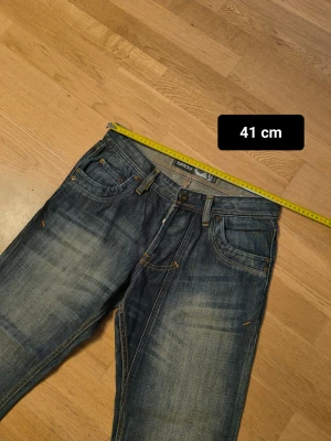 Blå jeansbyxor med detaljerad bakficka - Snygga blå jeansbyxor från SMOG med coola slitningar och kontrastsömmar. Bakfickorna har ett mönstrat broderi som ger extra stil. Klassisk femficksmodell med normal passform och raka ben. Perfekt för dig som gillar en avslappnad look.