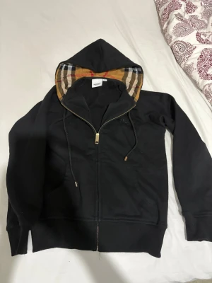 Svart hoodie från Burberry med dragkedja - Svart  hoodie från Burberry med klassiskt rutigt foder i huvan och broderad logga på bröstet. Tröjan har snörning i huvan, två fickor fram och dragkedja. Tillverkad i mjuk bomull med ribbade muddar och detaljer i elastan för extra komfort.
