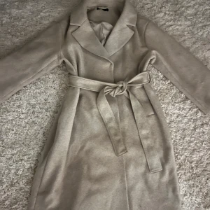 Beige kappa med bälte och krage - Snygg beige kappa med klassisk krage och knytbälte i midjan. Jackan har långärmade ärmar och en stilren, rak passform som ger en clean look. Perfekt för dig som gillar neutrala färger och vill ha en enkel men trendig jacka till höst och vinter.