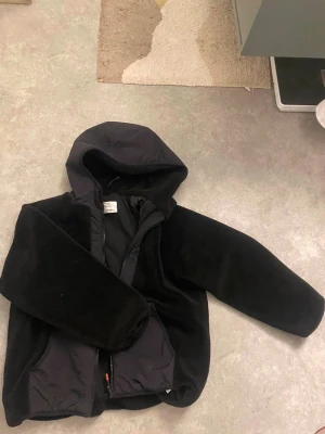 Svart teddyjacka med huva - Mysig svart teddyjacka med huva och dragkedja framtill. Jackan har två stora fickor och är tillverkad i mjukt fleece-material. Perfekt för kyliga dagar när du vill vara både varm och stilren. Storleken är 152cm men är lite större än så😊 Köpt ny från Zara 