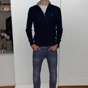 Marinblå merinoulltröja Morris S - Snygg marinblå stickad tröja från Morris i 100% merinoull. Tröjan har half zip med silverfärgad dragkedja och en liten broderad logga på bröstet. Kragen är beige på insidan och modellen är långärmad med ribbade muddar. Perfekt för dig som gillar stilrena och klassiska plagg. Använd fåtal gånger och priset är inte hugget i sten👍