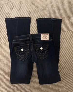 True Religion Becca Bootcut Jeans, strl 27/S - Mörkblå jeans från True Religion, modell Becca med bootcut och lowwaist. Snygga kontrastsömmar och ikoniska fickdetaljer med vita knappar bak. Klassisk jeanslook i stretchigt bomullsmaterial, perfekt för dig som gillar en avslappnad men trendig stil. Inte super använda och är bra skick!🌸 Är i storlek 27 som motsvarar storlek S men kan även i vissa fall passa M