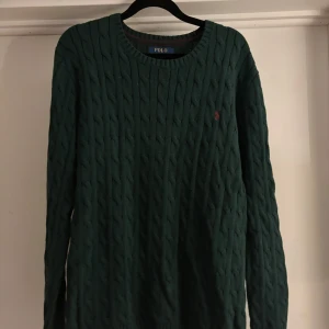 Mörkgrön Cable-Knit Polo Ralph Lauren - Mörkgrön kabelstickad tröja från Polo Ralph Lauren i storlek M. Klassisk rund halsringning och långärmad modell med broderad röd logga på bröstet. Perfekt för dig som gillar en tidlös och stilren look.