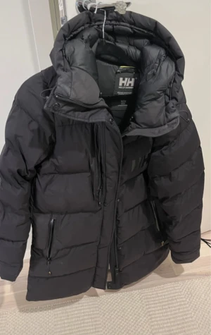 Svart pufferjacka från Helly Hansen - Säljer en svart pufferjacka från Helly Hansen med huva och snygga gulddetaljer på snörningarna. Jackan har dragkedja framtill, flera fickor och är perfekt för kalla dagar. Nypris 3499kr