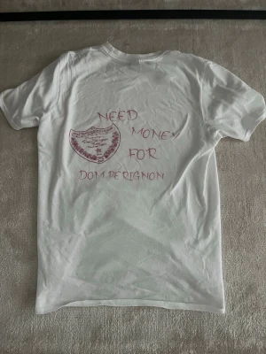 Vit t-shirt med Dom Pérignon-tryck - Vit t-shirt med korta ärmar och tryck i rosa på ryggen med texten 'Need money for Dom Pérignon' och en Dom Pérignon-logga. Skön passform och tillverkad i mjuk bomull, perfekt för dig som gillar statement-plagg och champagne vibes.