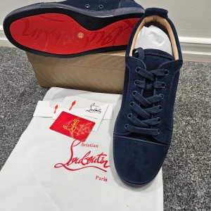 Mörkblå sneakers från Christian Louboutin - Snygga mörkblå sneakers från Christian Louboutin med lyxig mocka och klassisk röd sula. Skorna har rund tå, platt sula och snörning framtill. Insidan är beige och de har diskreta detaljer som ger en exklusiv vibe. Perfekt för dig som gillar statement-skor.