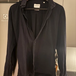 Mörkblå skjorta från Lindbergh - Mörkblå skjorta från Lindbergh i mjuk bomull. Klassisk krage och knappar framtill, med långärmad design. Perfekt för dig som gillar stilrena och enkla plagg. Skjortan har en normal passform och passar till många olika looks.