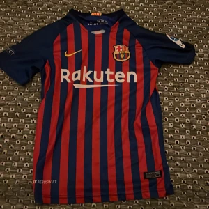 FC Barcelona Messi retro T-shirt  - Säljer en officiell FC Barcelona matchtröja från Nike med Messi 10-tryck på ryggen. Tröjan är blå och röd med vertikala ränder, korta ärmar och klubbmärke på bröstet. Materialet är lätt och andas, perfekt för fotboll eller samling.