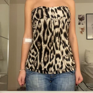 Leopardmönstrad tubtopp i beige och svart - Säljer en snygg tubtopp med leopardmönster i beige och svart. Toppen är axelbandslös och har en loose passform som sitter bekvämt över bröstet och midjan. Perfekt för dig som vill sticka ut med ett djuriskt print. Materialet känns mjukt och följsamt mot huden.