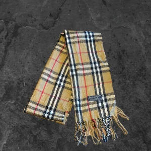 Burberry halsduk  - Klassisk rutig halsduk från Burberry i 100% lammull. Beige bas med svarta, vita och röda rutor, samt fransar i ändarna. Perfekt accessoar för att lyfta din outfit med en tidlös brittisk stil.