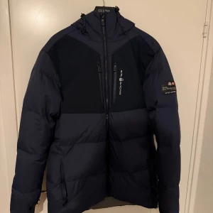 Mörkblå dunjacka Sail Racing - Säljer en mörkblå dunjacka från Sail Racing med hög krage, huva och flera dragkedjeförsedda fickor. Jackan har logga på bröstet och är riktigt varm och skön för kalla dagar. Perfekt för dig som vill ha en stilren och sportig look.