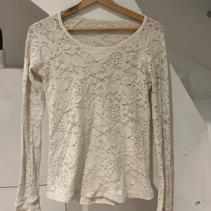 Vit spetsblus med lång ärm - Superfin vit blus i spets med lång ärm och rund halsringning. Blusen har ett vackert blommigt hålmönster över hela plagget och är lätt transparent.