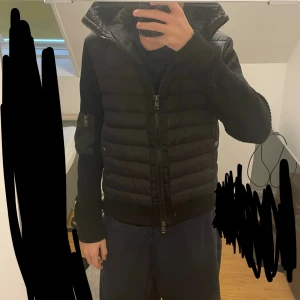 Svart Moncler Cardigan  - Säljer en svart pufferjacka från Moncler med stickade ärmar och huva. Jackan har dragkedja framtill, ribbade muddar och en ficka med dragkedja på ärmen. Klassisk Moncler-logga på vänster ärm. Perfekt för dig som vill ha en snygg och varm jacka med streetkänsla.