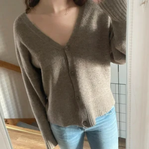 Beige stickad kofta med v-ringning - Mysig beige stickad kofta med djup v-ringning och knappar framtill. Koftan har långa ärmar med breda ribbade muddar och en avslappnad passform. Perfekt att slänga på sig över en t-shirt eller linne för en chill look.