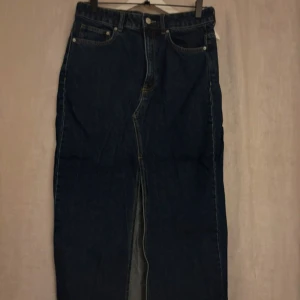 Mörkblå jeanskjol från H&M - Säljer en mörkblå lång jeanskjol från H&M med klassisk femficksdesign och slits framtill. Kjolen har hög midja och raka linjer, perfekt för en cool och avslappnad look. Gjord i kraftig denim med snygga kontrastsömmar.