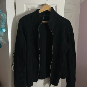 Svart stickad cardigan jacka 157 - Svart stickad cardigan jacka från 157 med grov ribbad struktur och dragkedja framtill. Jackan har hög krage och långärmade ärmar, perfekt för lager på lager. Enkel och stilren design som funkar till många olika looks. Aldrig använd och hänger bara inne i min garderob ‼️ Super snygg som en kofta till en kall sommardag eller bara under våren eller hösten/vintern 🥰☺️❤️