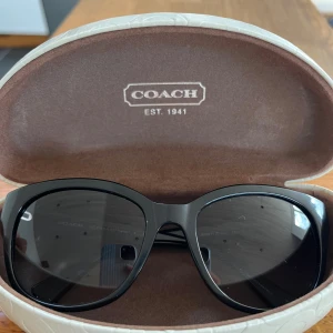 Svarta solglasögon från Coach - Snygga svarta solglasögon från Coach med bred båge och diskret logga på sidorna. Bågarna har ett präglat mönster och glasen är mörkt tonade. Kommer med fodral i vitt med Coach-logga. 