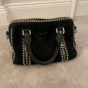 Svart handväska Zadig & Voltaire mocka - Cool svart handväska i mocka från Zadig & Voltaire med silverfärgade nitar och dragkedjor. Väskan har två handtag och avtagbar axelrem med logga. Snygga detaljer och rockig vibe, perfekt för dig som gillar statement-accessoarer.