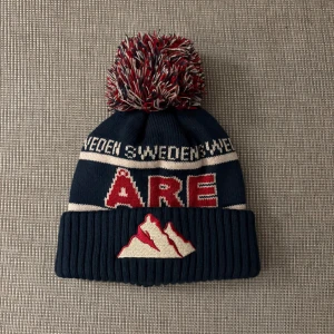 Mörkblå Åre mössa med tofs - Säljer en mörkblå stickad mössa från Åre med stor röd, vit och blå tofs. Mössan har broderad bergsdetalj och texten 'SWEDEN' samt 'ÅRE' i rött och vitt. Ribbad uppvikning och klassisk vinterstil. Perfekt för kalla dagar och fjällhäng. Den är aldrig andvänd.
