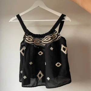 Svart broderad linnetopp från Zara - Svart linnetopp från Zara med breda axelband och broderade vita detaljer. Toppen har knytning i ryggen och är tillverkad i ett lätt material som passar perfekt för varma dagar. Helt ny och oanvänd med lappen kvar.